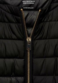 Gros plan d'une veste noire matelassée avec une fermeture éclair en métal doré partiellement ouverte et une étiquette indiquant "Imperméable, Fabriqué au Cambodge".