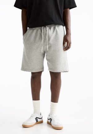 Mężczyzna ubrany w jasnoszare szorty ze ściągaczem, białe skarpetki, białe buty sportowe z czarnymi paskami i czarną koszulkę, stojący na białym tle.