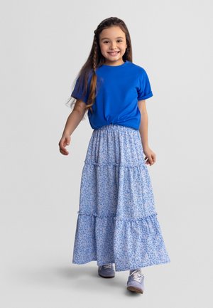 Jeune fille souriante aux longs cheveux, portant un t-shirt bleu, une longue jupe fleurie et des baskets violettes, marchant vers l'avant sur un fond uni.