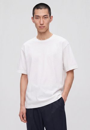 Jeune homme aux cheveux courts portant un t-shirt blanc uni et un pantalon foncé, debout avec une main dans la poche devant un fond clair.