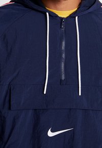 Marinblå vindjacka med huva, vit dragkedja och snoddar. Har en framficka och en vit Nike-logotyp längst ner fram.