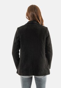 Donna con lunghi capelli castani, che indossa un blazer di velluto a coste nero e jeans azzurri chiari, girata di spalle su uno sfondo bianco uniforme.