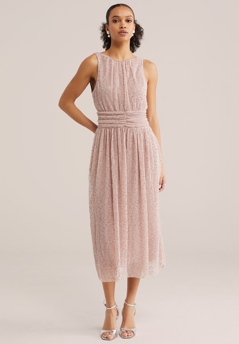 Kobieta w różowej, bezrękawnikowej sukience midi z cekinami i marszczonym pasem, w srebrnych szpilkach z paskiem wokół kostki, stojąca na neutralnym tle.