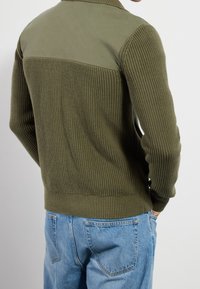 Olivgrön stickad tröja med ett mjukt tygpanel på axlarna, ribbad textur, åtsittande ärmslut och rak fåll.