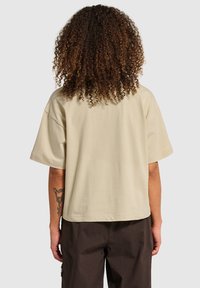 Beige oversized t-shirt met korte mouwen, gemaakt van zachte stof. Heeft een rechte zoom en een ontspannen pasvorm. Zichtbare tatoeage op de arm.