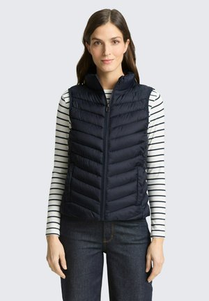 Femme portant une veste matelassée bleu marine sur une chemise blanche à manches longues avec des rayures horizontales noires, associée à un jean foncé, debout et face à l'avant.