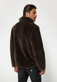 Giacca in pile marrone con colletto alto, caratterizzata da una vestibilità rilassata e una texture morbida. La vista posteriore mostra le maniche che arrivano ai polsi.