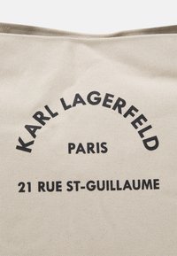 Beżowa torba na ramię z płótna, z czarnym napisem: "KARL LAGERFELD", "PARYŻ" i "21 RUE ST-GUILLAUME". Prosty design z wytrzymałą strukturą.