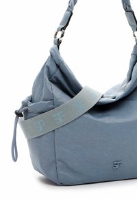 Sac à main bleu en matériau souple et texturé. Dispose d'une bandoulière tressée et d'une bande avec logo, avec une fermeture à cordon en haut.