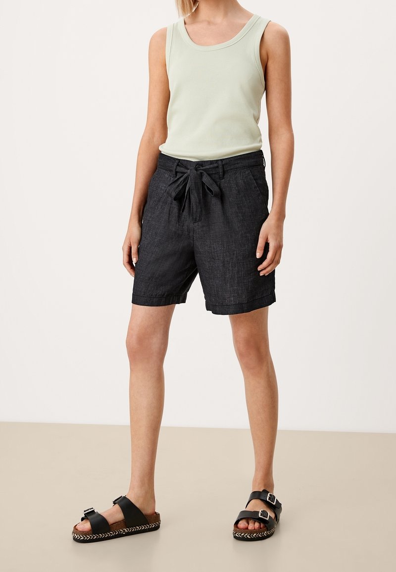 Femme portant un haut sans manches vert clair, un short noir avec taille à nouer et des sandales noires à boucle, debout sur un sol et un fond neutres.