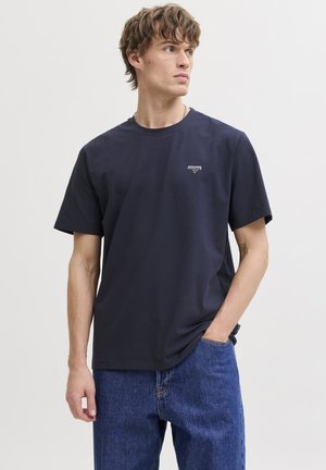 Joven con camiseta azul marino con pequeño logo blanco en el pecho y vaqueros azules, de pie con una mano en el bolsillo, mirando hacia un lado.