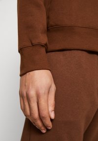 Brown sweatshirt fabric featuring a smooth texture with ribbed cuff detail, paired with matching brown pants. Hand resting on fabric.