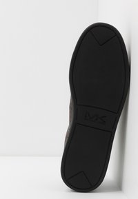 Svart gummisula på en sko med texturerad yta, prydd med logotypen "MK" präglad. Sidan av skon är klädd med mönstrad tyg i dämpade toner.