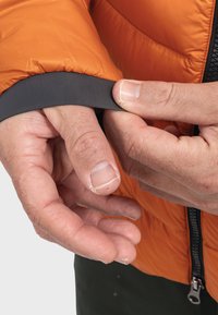 Oranger isolierter Jackenärmel mit einem grauen gerippten Bündchen. Hände, die das Bündchen festhalten und die Textur sowie Elastizität der Materialien zeigen.