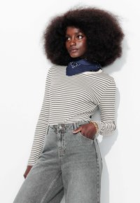 Haut à manches longues rayé en crème et noir, associé à un jean en denim gris taille haute. Présente une écharpe bleue et des cheveux texturés et volumineux.