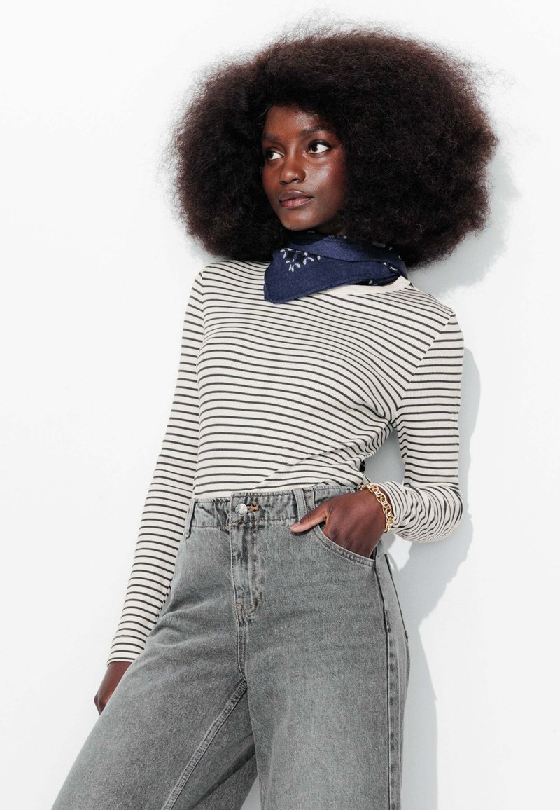 Haut à manches longues rayé en crème et noir, associé à un jean en denim gris taille haute. Présente une écharpe bleue et des cheveux texturés et volumineux.