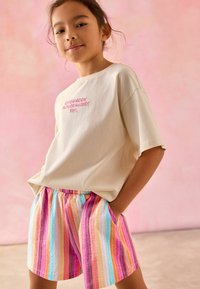 T-shirt beige ample avec un texte brodé. Shorts colorés à rayures avec du rose, de l'orange, du bleu et du jaune, en tissu léger.