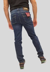Uomo che indossa jeans blu scuro con etichette nelle tasche posteriori, una maglietta nera e sneakers multicolori, in piedi davanti a uno sfondo grigio uniforme.