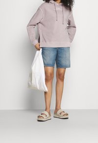 Svetle fialová mikina s kapsou na přední straně a šňůrkou, spárovaná s modrými džíny krátkými kalhotami a béžovými platformovými sandály s metalickým akcentem.