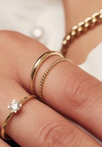 Three gold rings on a hand: a smooth band, a beaded band, and a gem-set band featuring a round clear stone, all with a polished finish.