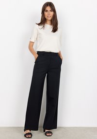 Pantalons noirs à jambes larges avec une texture lisse et une coupe ajustée, accompagnés d'un haut à manches courtes beige clair. Sandales noires à brides.