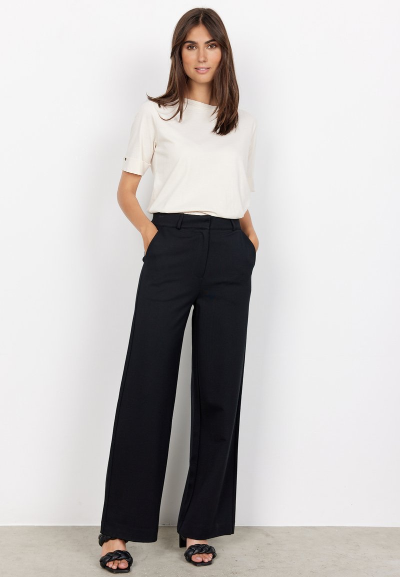 Pantalons noirs à jambes larges avec une texture lisse et une coupe ajustée, accompagnés d'un haut à manches courtes beige clair. Sandales noires à brides.