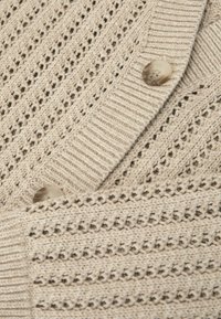 Beige knitted cardigan with openwork pattern, ribbed edges, and two marbled beige buttons fastened along the front.