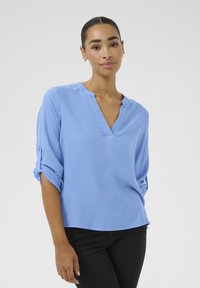 Blouse bleu clair à texture gaufrée avec un décolleté en V et des manches retroussées. Pantalon noir visible, mannequin face à un fond neutre.