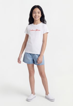 Χαμογελαστή κοπέλα με μακριά σκούρα μαλλιά, που φοράει λευκό t-shirt Calvin Klein, ανοιχτόχρωμα τζιν σορτς και λευκά slip-on sneakers, στέκεται σε λευκό φόντο.