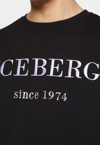 T-shirt in cotone nero con testo ricamato bianco in evidenza: "ICEBERG" e "dal 1974". Collo rotondo, texture liscia.