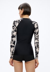 Moteris su trumpais tamsiais plaukais, matoma iš nugaros, dėvi juodą aptemptą ilgomis rankovėmis viršutinį drabužį su baltomis abstrakčiomis raštuotomis rankovėmis ir juodus šortus.