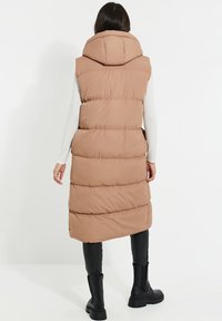 Veste longue matelassée de couleur marron clair avec capuche, présentant des coutures horizontales et une coupe décontractée. Associée à un pantalon noir ajusté et des bottes.