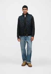 Tmavě modrá prošívaná vesta s vysokým límcem přes tmavě šedý svetr, doplněná o světle modré džínové kalhoty a světle hnědé slip-on boty.