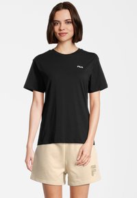 T-shirt in cotone nero con collo a crew e maniche corte, con un piccolo logo FILA bianco sul lato sinistro del petto. Abbinato a pantaloni corti beige.