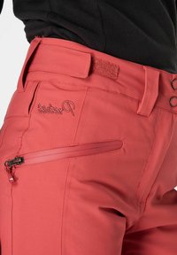 Pantaloni da esterno rosa con una texture liscia, dotati di tasca con zip e logo ricamato. Visibili passanti per cintura e dettaglio con chiusura a strappo.