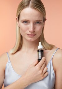 Braune Glas-Tropfflasche mit weißem Deckel, beschriftet mit "Garnier AHA+BHA Serum" in grüner und schwarzer Schrift. Eine Person hält sie vor einem hellblauen Stoffoberteil.
