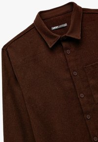 Camisa marrón de botones hecha de una tela texturizada. Presenta un cuello clásico, un bolsillo frontal y cinco botones marrones a lo largo de la tapeta.