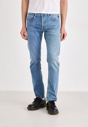 Homme portant un jean slim bleu clair et des chaussures noires à lacets, debout sur un fond uni.