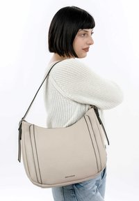 Sac à bandoulière en cuir beige avec une forme courbée, surface texturée, et une sangle en accent noir. Présente des fermetures éclair des deux côtés.