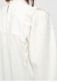 Blusa bianca e testurizzata con dettagli a ruches lungo le maniche e il collo, e chiusura con bottoni sul retro.