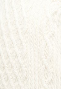 Gros plan sur un tissu tricoté blanc avec un motif en relief de torsades composées de mailles verticales et diagonales entrelacées.