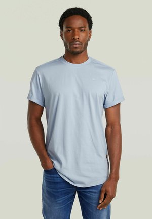 Uomo con capelli corti e ricci che indossa una maglietta azzurro chiaro con maniche arrotolate e jeans blu, in piedi contro uno sfondo semplice.