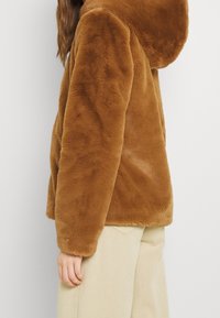 Veste en fausse fourrure marron avec une capuche, présentant une texture douce et une coupe décontractée, associée à un pantalon large de couleur claire.