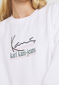 Sudadera blanca con el logo de texto en negro y verde "Kani" con líneas de acento rosas debajo. Tejido suave con cuello y puños de canalé.