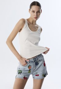 Top in cotone bianco a costine abbinato a shorts di jeans leggeri blu chiaro con ricami di frutta colorata. Il design include spacchi laterali e una forma aderente.
