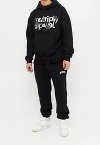 Sudadera negra con texto impreso en verde y blanco, bolsillo frontal, combinada con pantalones de chándal negros que tienen un pequeño logo, usados con zapatillas blancas.