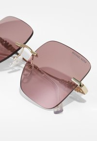 Óculos de sol quadrados com lentes cor-de-rosa, detalhes metálicos dourados e pontas de templos transparentes. Logo visível na lente, design moderno e elegante.