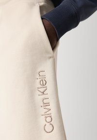 Beige jogger pants with a ribbed navy cuff, featuring "Calvin Klein" embroidered in light brown on the side. Soft fabric texture.
