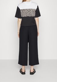 Mujer vestida con pantalones anchos negros y una blusa de manga corta en negro, blanco y estampado de leopardo, de pie descalza sobre un fondo liso.
