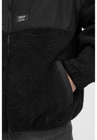 Giacca in pile nera con una parte inferiore morbida e testurizzata e una parte superiore in tessuto liscio. Presenta una chiusura con zip e tasche laterali. Etichetta sul davanti.